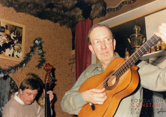 Constant playing guitar with other man in the background